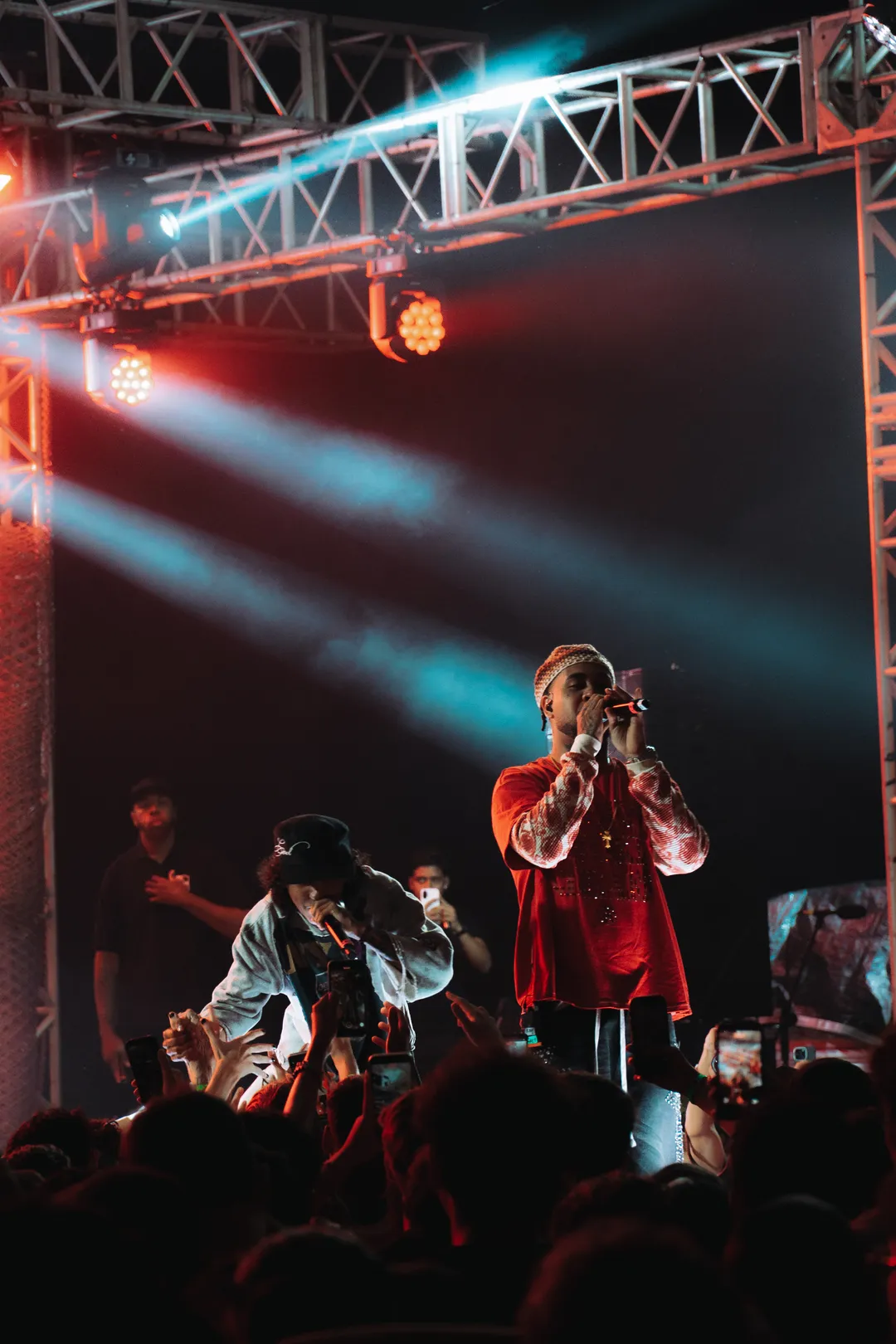 KAWE cantando sob refletores vermelhos, com outra pessoa no palco ao lado